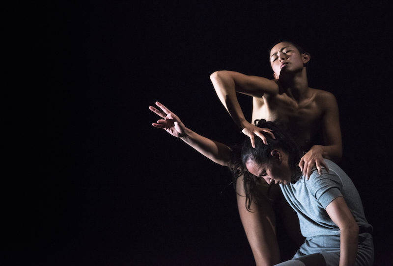 A naked dancer presses her hands atop another performer's head and shoulder.